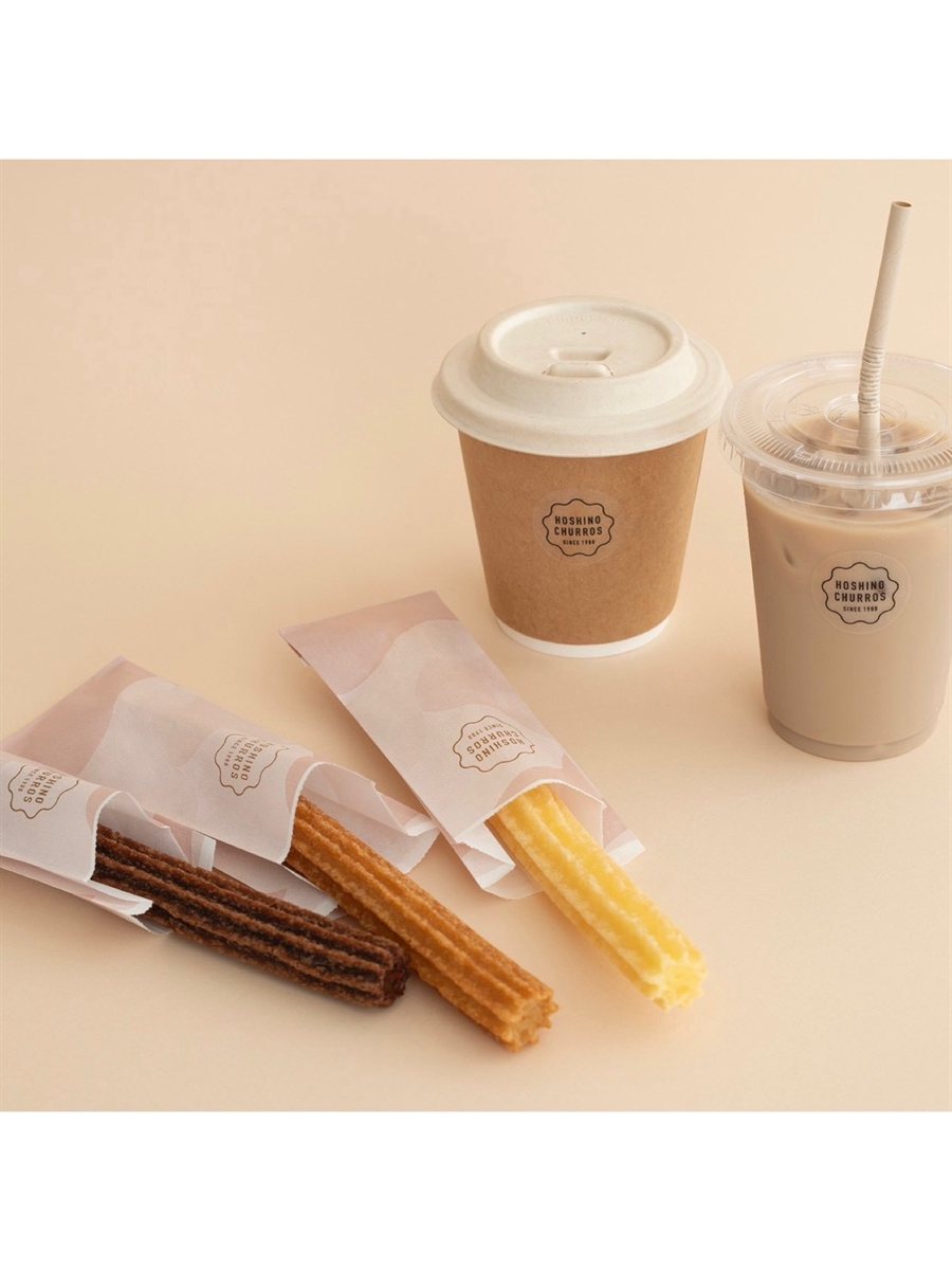 ベージュ背景に並ぶ〈HOSHINO CHURROS〉のチュロス3種（プレーン・シナモン・チョコレート）と、HOSHINO CHURROSロゴ入りのホットコーヒー・アイスドリンク。薄ピンクのチュロス袋にすっと差し込まれた一本