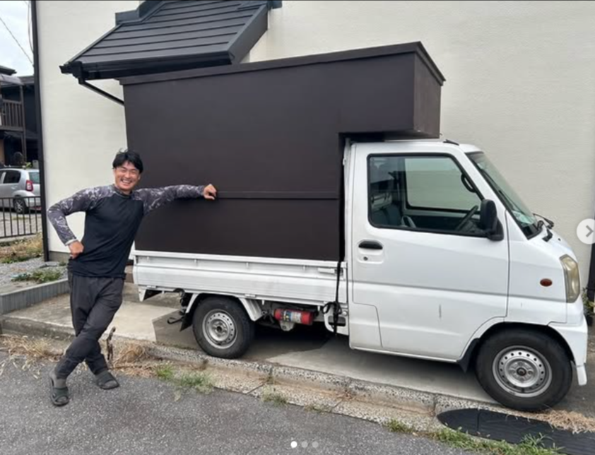 白い軽トラックの荷台に焦げ茶色の木の箱を載せた自作キッチンカーと、車体に手をかける店主