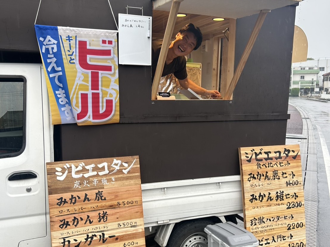 販売窓から顔を出す店主と、車体の前に並ぶ2枚の手書きメニュー看板